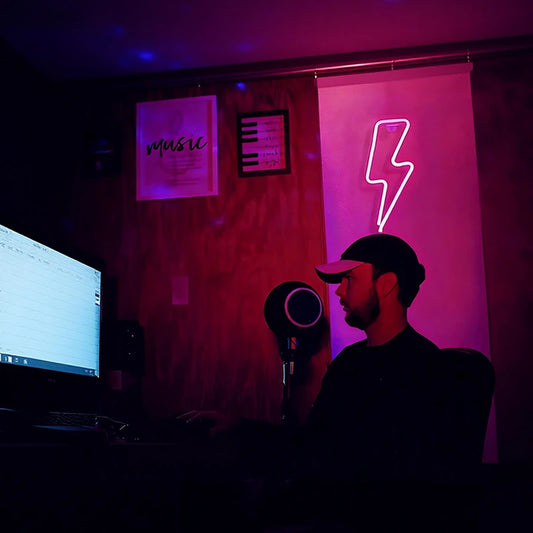 Person in a dark room with a computer and microphone, illuminated by hot pink lightning bolt neon light.
