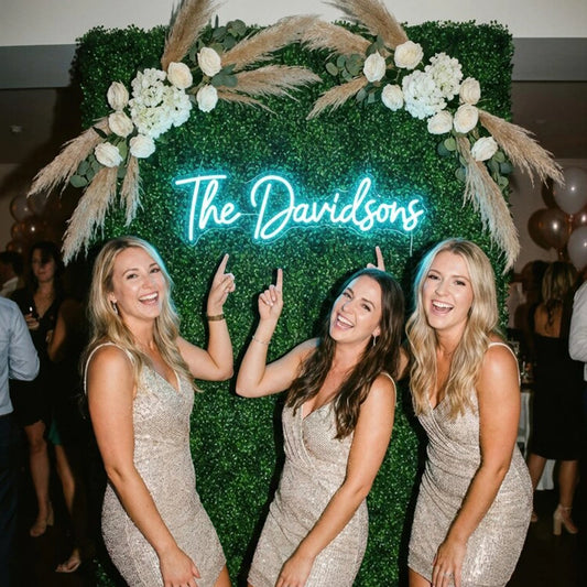 Three women pointing at a neon sign with letters 'The Davidsons' in a wedding scene