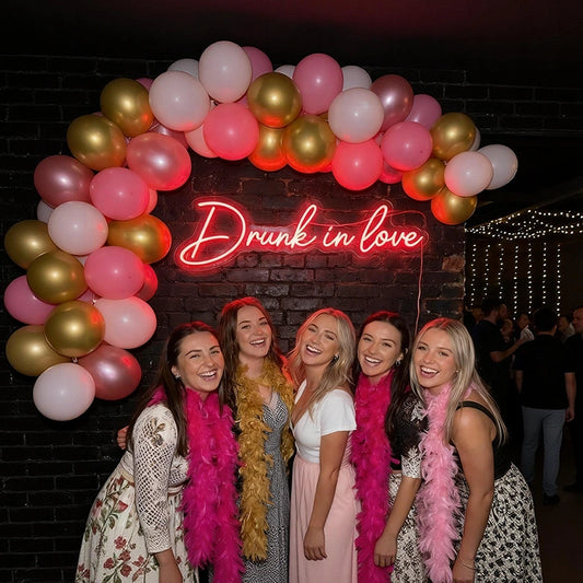 Five women smiling and posing together in front of a black brick wall decorated with a pink and gold balloon arch and a glowing neon sign that reads 'Drunk in love'. Each woman is wearing a bright pink feather boa, and the women are wearing dresses in white, pink, and black.