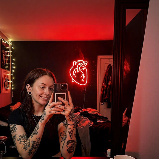 Person with tattoos using a phone in a room with a heart-shaped neon light.