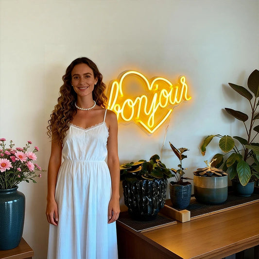 Woman in a white dress standing next to a neon 'bonjour' sign with plants in the background.