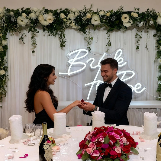 Man and woman at a wedding table with floral decorations and 'Bride to Be' sign.