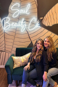 Two women sitting on a couch in front of a decorative wall with 'Soul Beauty Co' neon sign.