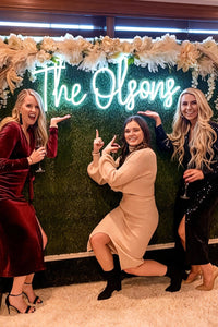 Three women posing in front of a decorative wall with 'The Olsons' neon sign.