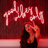 Woman making a peace sign in front of a neon sign with 'good vibes only' text.