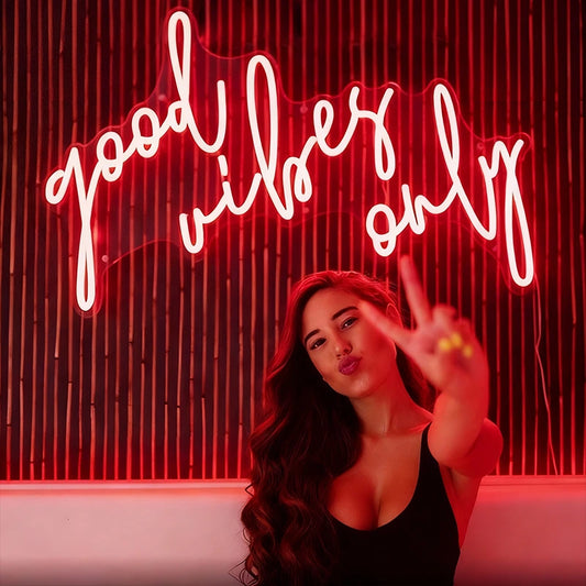 Woman making a peace sign in front of a neon sign with 'good vibes only' text.