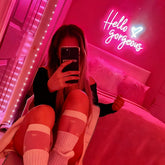 Person taking a mirror selfie in a room with pink lighting and 'Hello Gorgeous' neon sign.