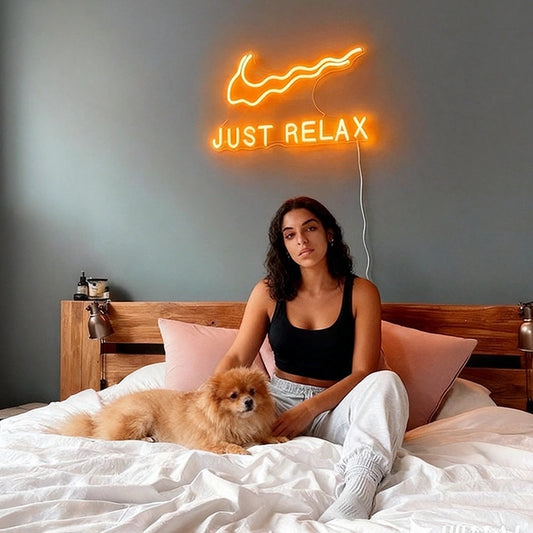 Woman sitting on a bed with a dog, neon sign above reading 'Just Relax' in a bedroom setting.