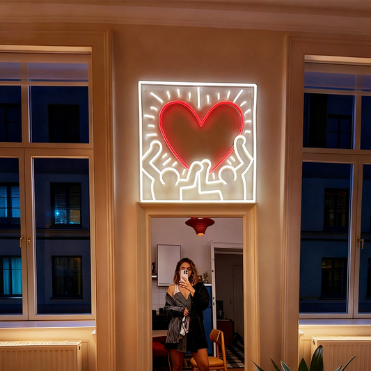 A square illuminated wall sign featuring a glowing red heart and white outline figures inspired by Keith Haring, mounted above a doorway in a warmly lit interior. A person is visible taking a mirror selfie in the reflected room below.