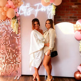 Two women posing in front of a 'Let's Party' sign with balloons and a sequin curtain.