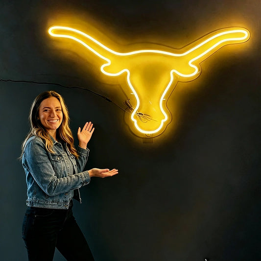A woman in a denim jacket stands beside a glowing lomon yellow neon sign shaped like a longhorn steer head on a dark wall.