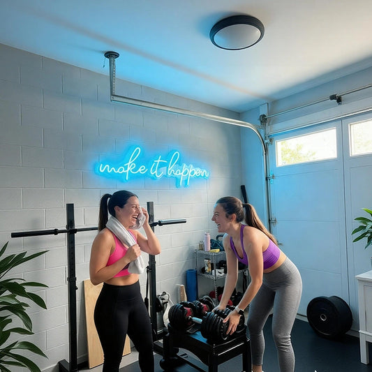 Two women in athletic wear inside a home gym with a glowing ice blue neon sign that reads 'make it happen' on a light gray brick wall. One woman stands on the left in a pink sports bra and black leggings, while the other bends over a weight bench holding a barbell on the right in a purple sports bra and gray leggings.