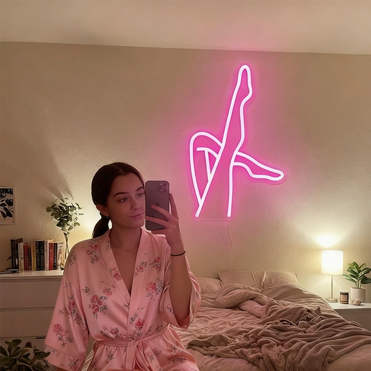 A woman in a floral satin robe takes a mirror selfie in a bedroom, with a glowing hot pink neon wall sign depicting a stylized female sexy legs behind her.