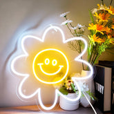 Neon light shaped like a sun with a smiley face, placed on a surface with flowers and books in the background.