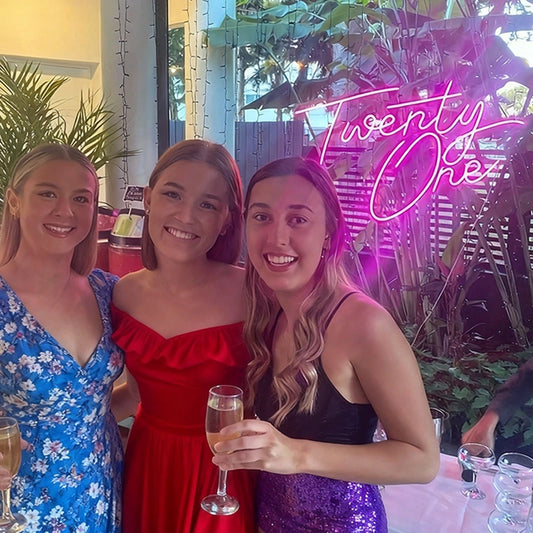 Three women posing together with drinks in a festive setting, featuring a ' Twenty One' neon birthday sign in the background.