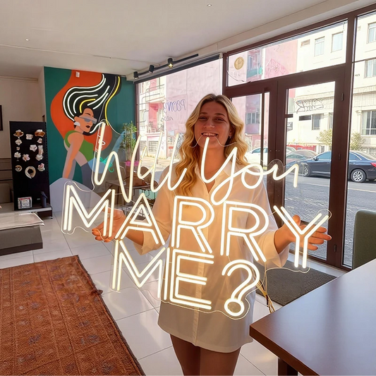Woman holding a neon sign asking 'Will you marry me?' in a room with large windows.