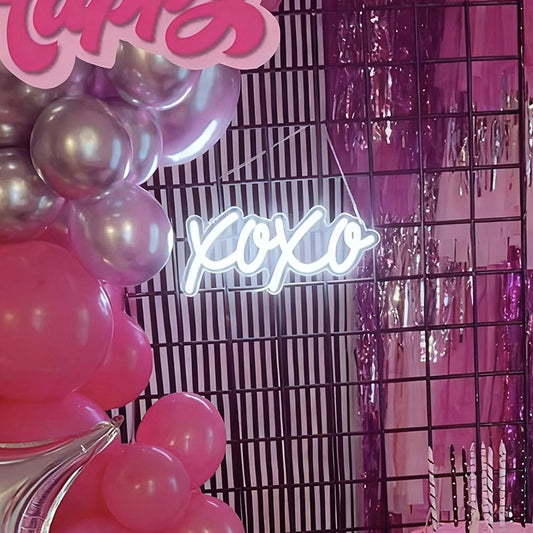 Pink and silver balloon garland, a black wire grid backdrop, and a glowing white 'xo xo' neon sign against a pink tinsel background.