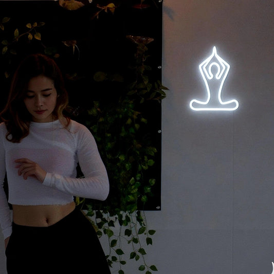 Woman standing next to a neon yoga pose sign on a wall with greenery.