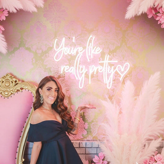 A woman in a navy off-the-shoulder dress stands beside a pink ornate chair against a pink floral backdrop with a glowing white script sign reading 'You're like really pretty' and pampas grass decorations.