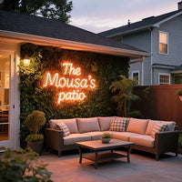 Outdoor patio scene at dusk with a wicker sectional sofa, coffee table, hedge wall, and a glowing neon-style sign reading “The Mousa’s patio.”.