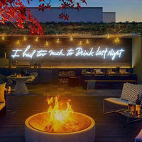 A rooftop bar or lounge at dusk with a glowing white neon sign reading 'I had too much to Drink last Night' mounted on a wood wall. A round fire pit burns in the foreground, with patio seating, tables, greenery, and red autumn leaves visible above.