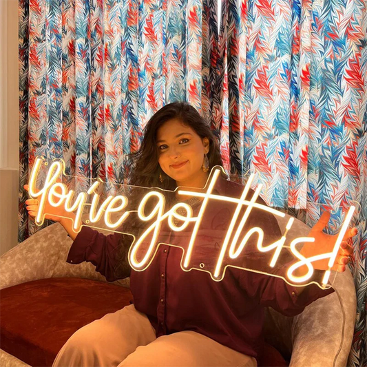 Person sitting on a couch holding a neon sign with 'You've got this!' text in front of colorful curtains.