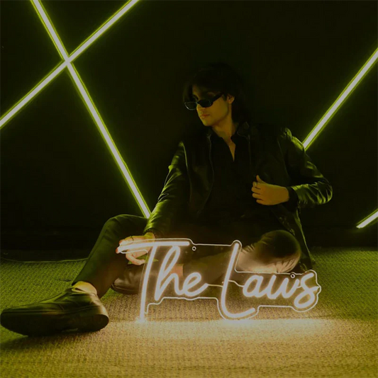 Person sitting on the floor with a neon sign displaying 'The Laws' in a dark room with geometric light patterns.
