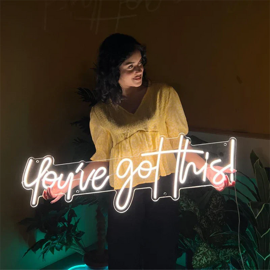 Woman holding a neon sign with 'You've got this!' text in a dimly lit room.