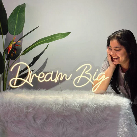 Person sitting on a white surface with a 'Dream Big' neon sign and plants in the background