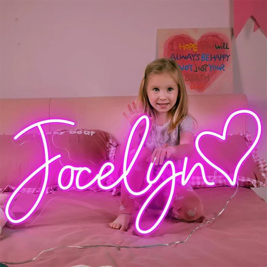 Young girl sitting on a bed with pink bedding and neon signs, including one with 'Jocelyn' and a heart.