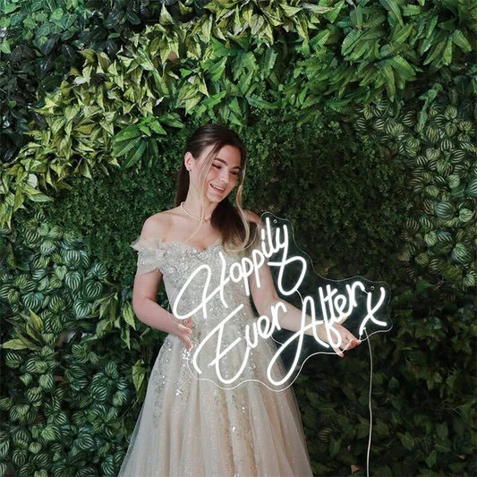 Woman in a beige off-shoulder gown holding a 'Happily Ever After' sign against a green leafy background