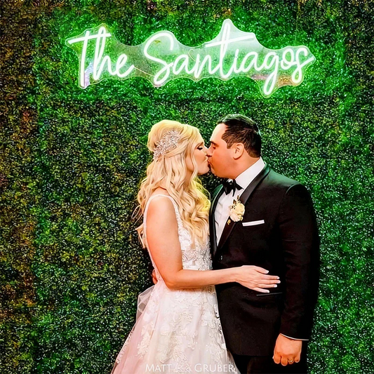 Couple in formal attire kissing in front of a green leafy background with 'The Santigas' neon sign.