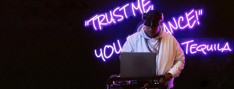 A man playing a DJ music device under a black background with purple neon sign quote behind