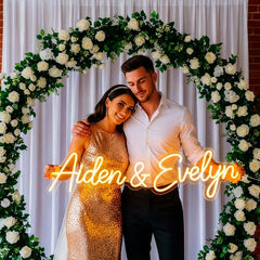 Couple standing under a floral arch with 'Aiden & Evelyn' neon sign