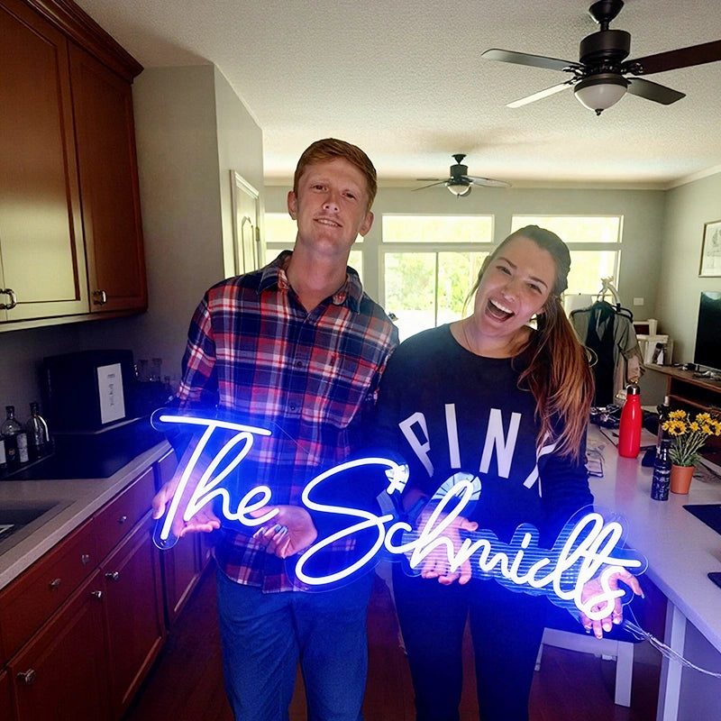 A man and a woman holding a blue neon sign together that reads 'The Schmilts' in a house.