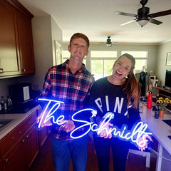 A man and a woman holding a blue neon sign together that reads 'The Schmilts' in a house.