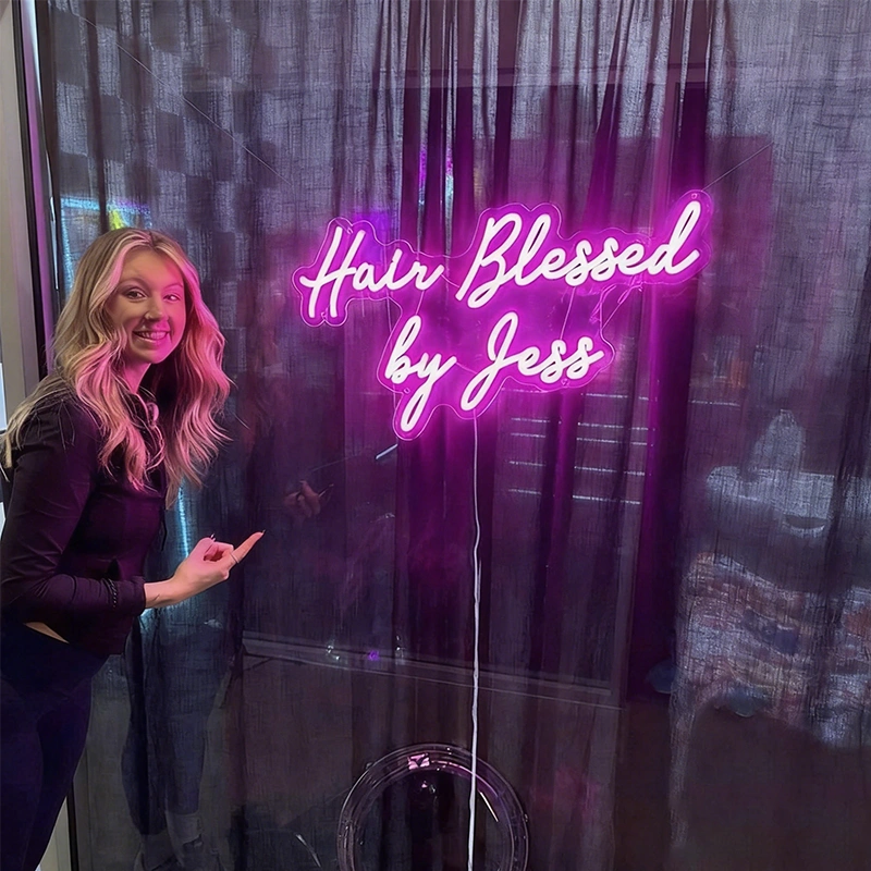 A curl hair woman pointing at a hot pink neon that reads 'The Hair Blessed by Jess' in a house.