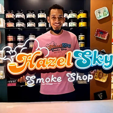 Person wearing a pink shirt with 'Hazel Sky Smoke Shop' logo neon sign in a store setting.