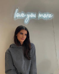 Woman standing in front of a neon sign that reads 'love you more' on a beige wall.