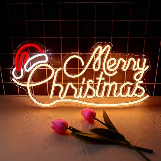 A glowing LED neon sign that reads 'Merry Christmas' with a red Santa hat accent, displayed on a wooden surface with two pink tulips in front against a dark grid background.