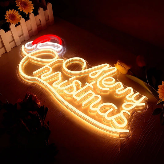 A glowing LED neon sign spelling out 'Merry Christmas' in warm white light, with a red and white Santa hat at the top, displayed on a dark surface with decorative flowers and a white picket fence in the background.