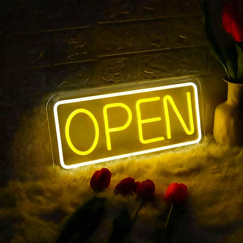 Neon 'OPEN' sign illuminated in a dimly lit setting with flowers around.