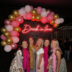 Five women smiling and posing together in front of a black brick wall decorated with a pink and gold balloon arch and a glowing neon sign that reads 'Drunk in love'. Each woman is wearing a bright pink feather boa, and the women are wearing dresses in white, pink, and black.