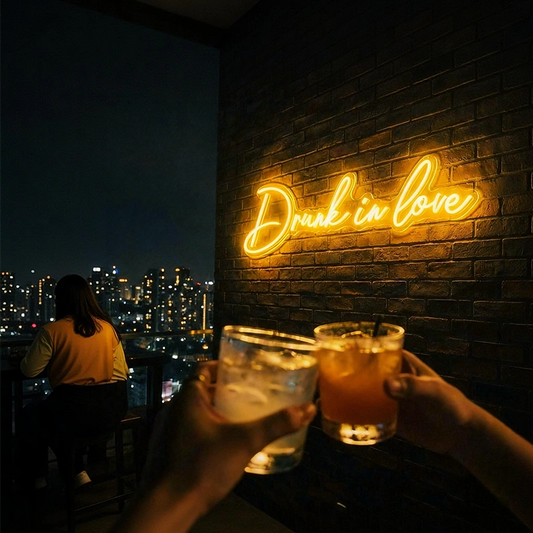 A glowing lemon yellow neon sign reading 'Drink in love' mounted on a dark brick wall at night, with two hands holding up glasses of iced drinks in the foreground and a person seated by the window in the background.