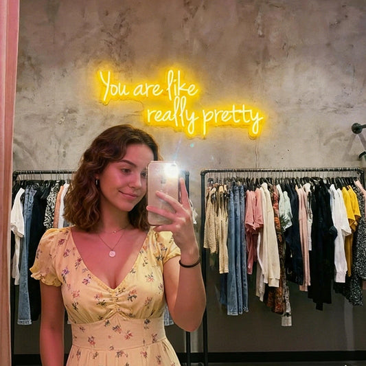 A woman takes a mirror selfie in a clothing boutique, wearing a pale yellow floral dress. Behind her is a gray concrete wall with a glowing lemon yellow neon sign that reads 'You are like really pretty' and racks of clothes.