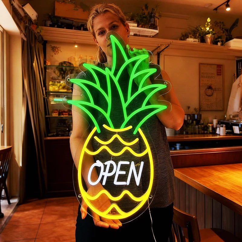 Person holding a neon pineapple sign with 'OPEN' text in a casual setting.