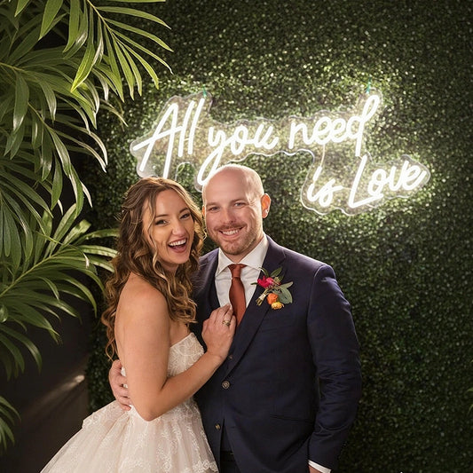 A smiling bride and groom posing together in front of a green leaf wall with a glowing white neon sign that reads 'All you need is Love'. The bride wears a strapless white lace wedding gown, and the groom is in a navy blue suit with a red tie and a floral boutonniere.