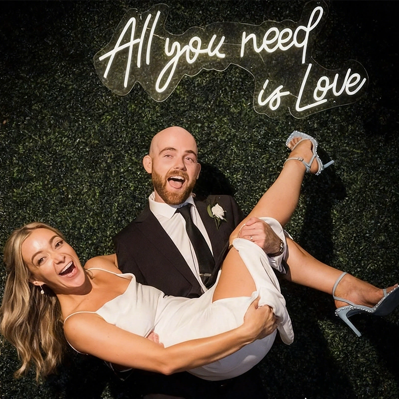 A smiling couple poses against a dark green hedge wall with a glowing neon sign that reads 'All you need is Love'. The man wears a black suit with a white shirt and black tie, while the woman wears a white sleeveless dress.