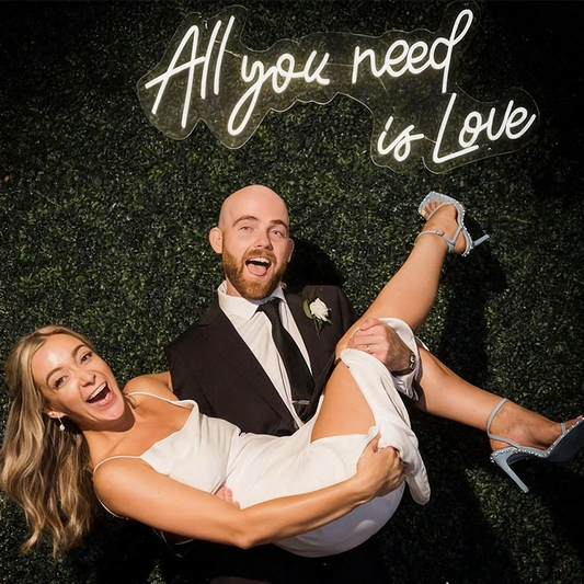 A smiling couple poses against a dark green hedge wall with a glowing neon sign that reads 'All you need is Love'. The man wears a black suit with a white shirt and black tie, while the woman wears a white sleeveless dress.