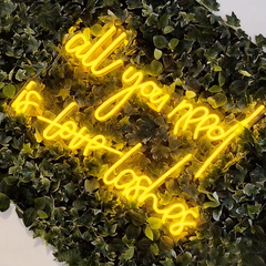 Neon sign with lemon yellow text 'all you need is love lashes' on a green leafy background